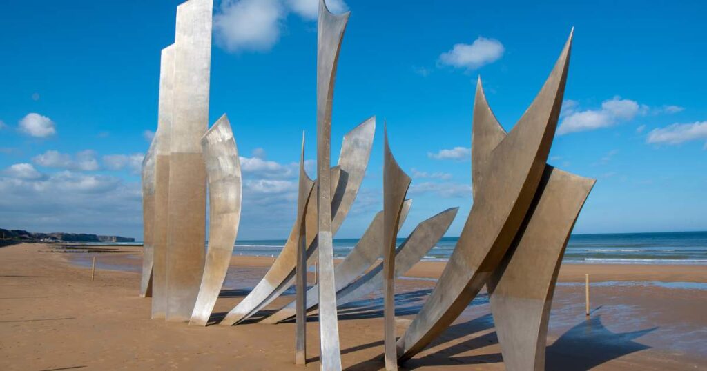 A massive metal sculpture standing on the sandy beach, overlooking the vast ocean in Normandy.